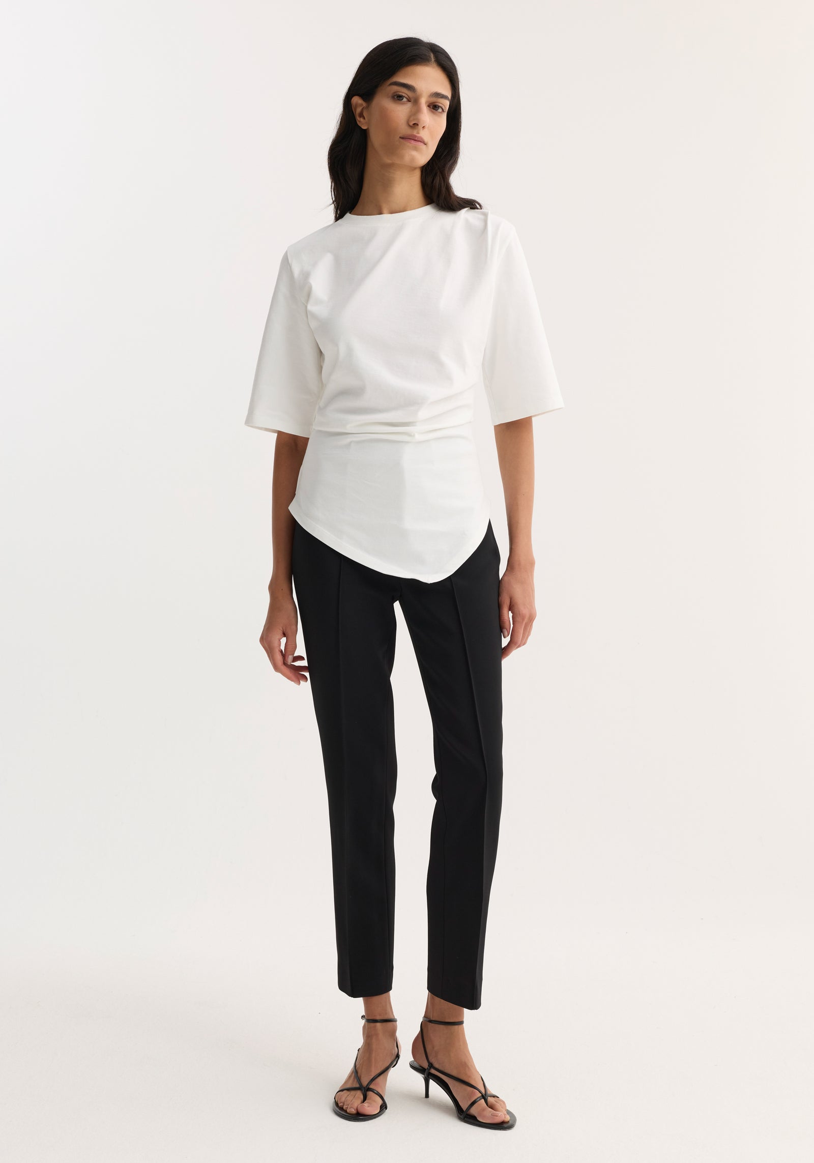 Woman in Reinvented Twisted T-Shirt | Optic White, featuring a sculptural asymmetrical drape and fitted waist, paired with slim-fit trousers and low-heel sandals.