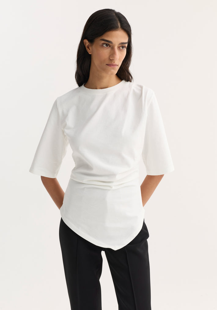 Reinvented twisted t-shirt in optic white, featuring an asymmetrical drape and fitted waist, worn with black trousers by a model with hands in pockets.