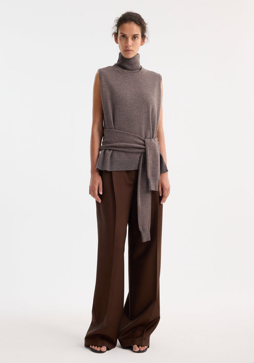 Model wearing Double-layered knit top | mud melange, featuring a sleeveless high-neck with waist tie, and paired with wide-leg chocolate trousers and black sandals.