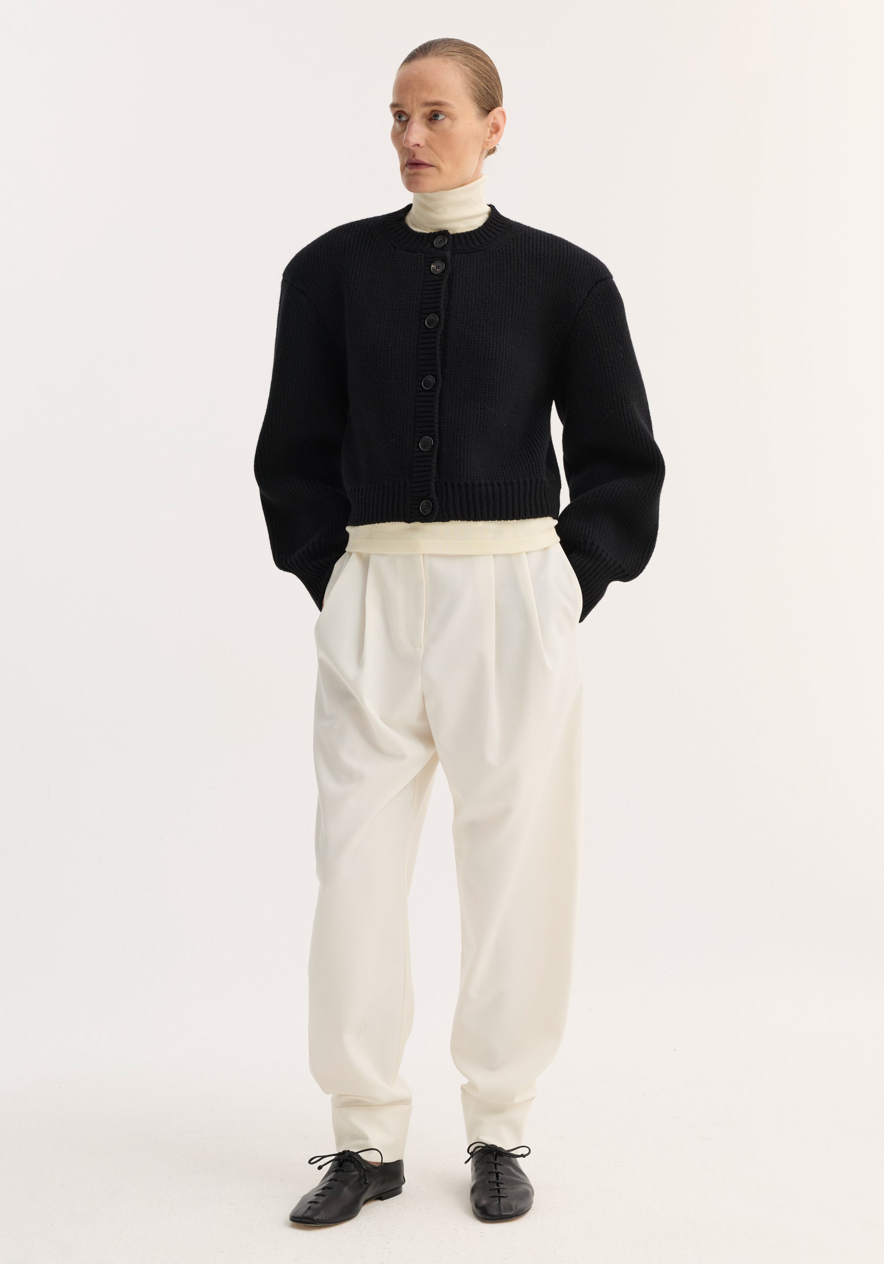 A model wearing a black Statement shoulder cardigan | black with oversized sleeves and a button-up front, paired with a white turtleneck, loose white pants, and black lace-up shoes.