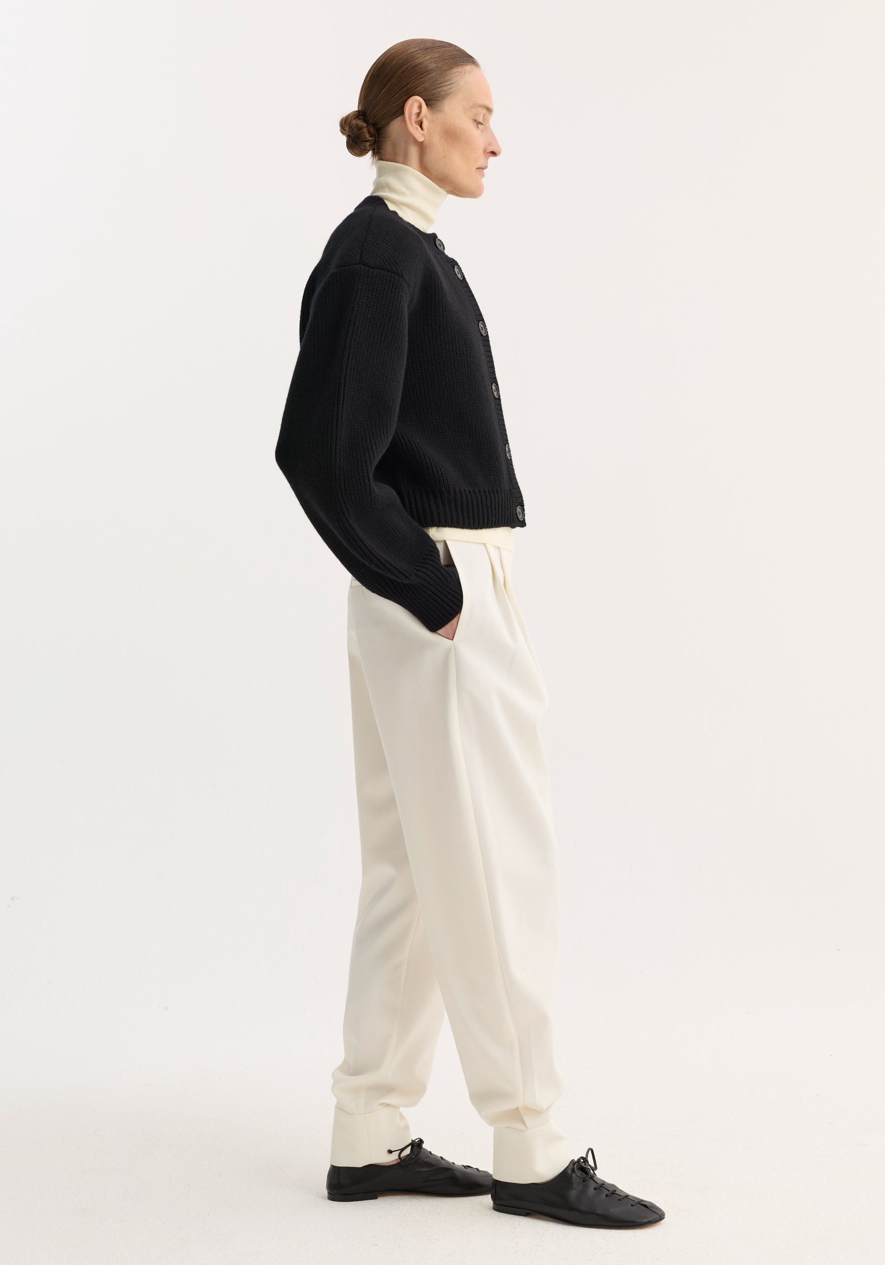 Woman with hair in bun wearing a black Statement shoulder cardigan | black over a white turtleneck, paired with loose white pants and black shoes, standing in profile against a plain background.