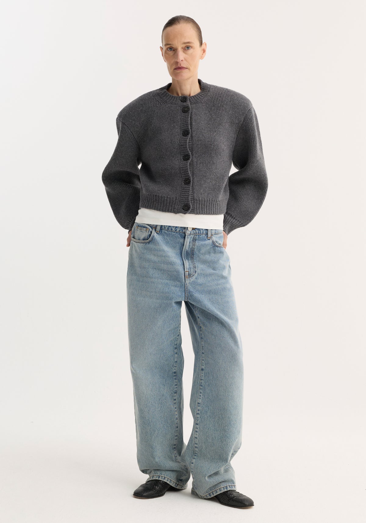 Model wearing a mid grey melange Statement shoulder cardigan with exaggerated shoulders, button-up front, ribbed cuffs and hem, paired with light blue wide-leg jeans and black shoes.