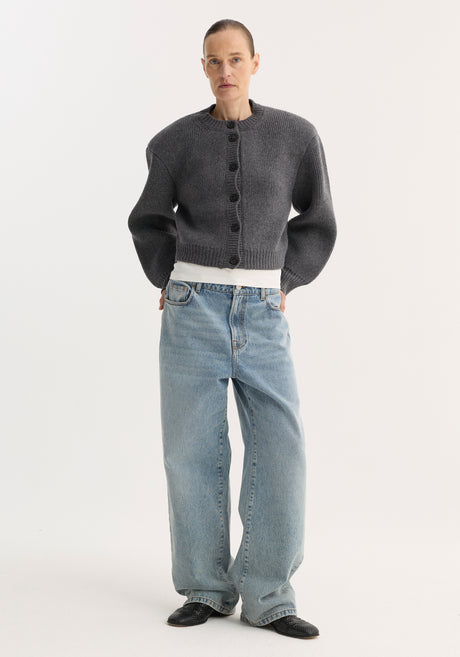 Person models the Statement Shoulder Cardigan in mid grey melange, featuring a structured oversized fit and button closure, paired with wide-leg jeans and pointed shoes.