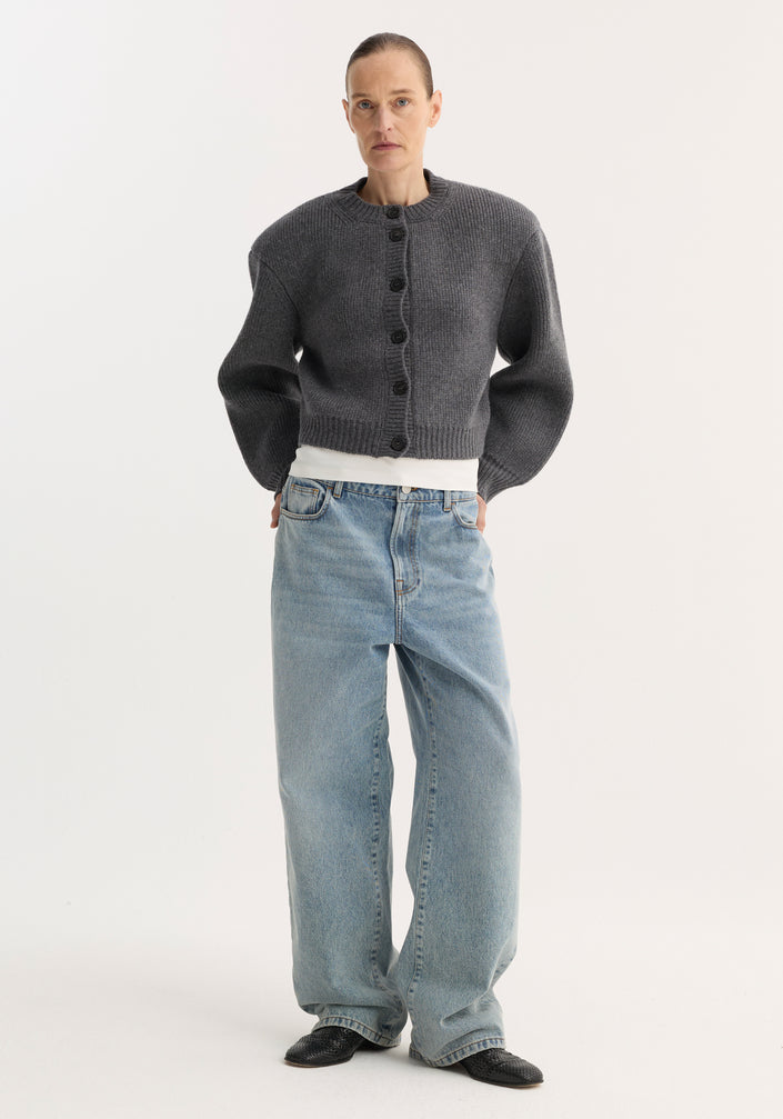 Person models the Statement Shoulder Cardigan in mid grey melange, featuring a structured oversized fit and button closure, paired with wide-leg jeans and pointed shoes.