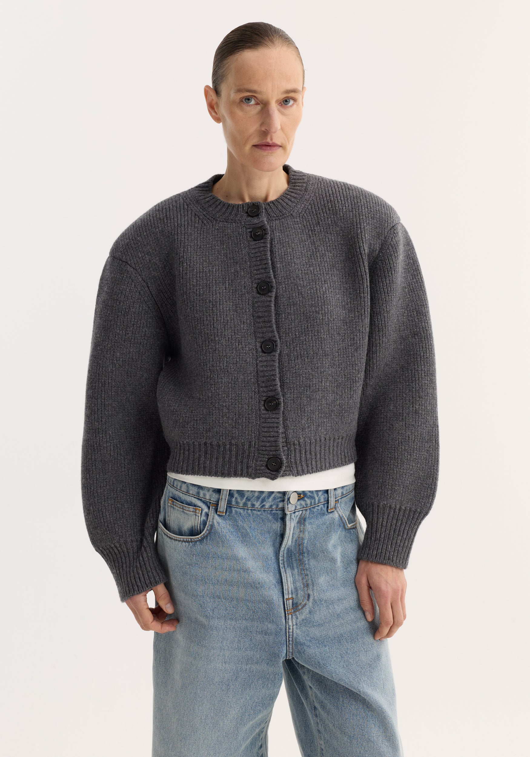 Woman wearing a dark grey chunky knit cardigan with statement shoulders and black buttons, paired with light blue jeans, styled as Statement shoulder cardigan | mid grey melange.