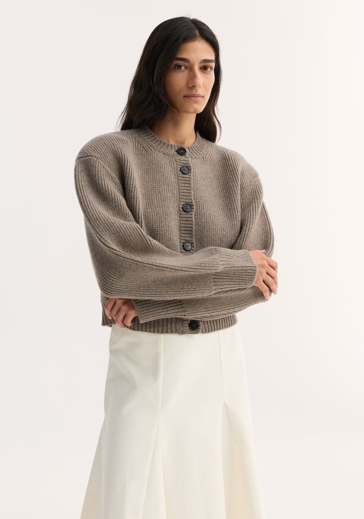 Person wearing the Statement Shoulder Cardigan in clay melange, featuring a chunky-knit with oversized buttons, sculptural shoulders, and a boxy fit over a high-waisted skirt.
