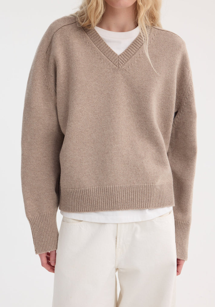 Person wearing a taupe melange Sleeve wrap jumper, featuring a V-neck and relaxed fit, layered over a white T-shirt, paired with cream pants.