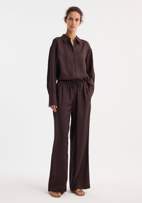 Model wearing wide leg silk trousers in mahogany, featuring an elasticated waistband and drawstring. Paired with a matching shirt and tan sandals.