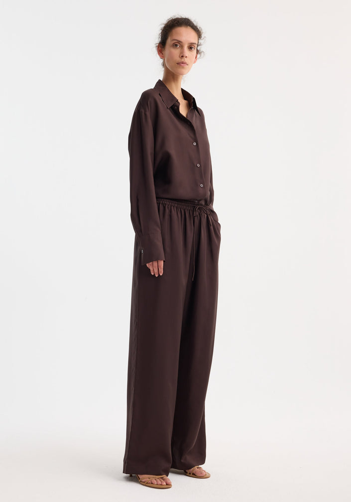 Model wearing wide leg silk trousers in mahogany, featuring an elasticated waistband and drawstring. Paired with a matching shirt and tan sandals.