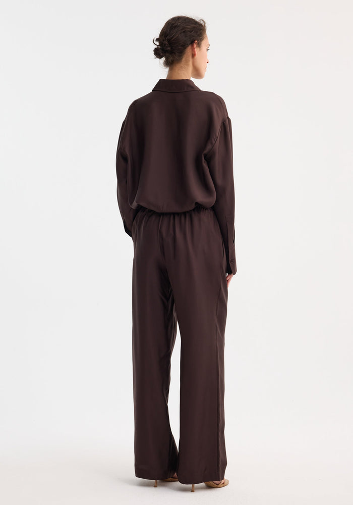 Woman wearing Wide leg silk trousers | mahogany, featuring an elastic waistband and drawstring, styled with a tucked blouse and light-tan heeled sandals.