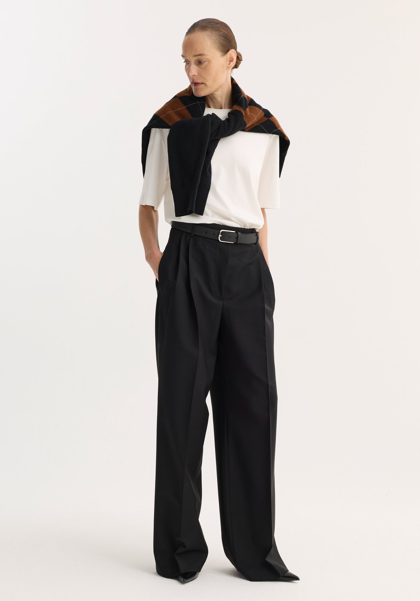 Model wearing Mid-waist tailored trousers | black, featuring a high-waisted, wide-leg cut with a black leather belt, paired with a white top and argyle sweater.