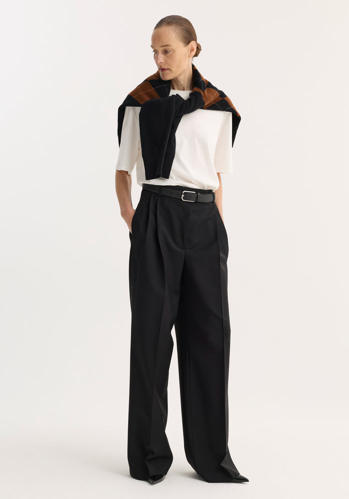 Model wearing Mid-waist tailored trousers | black, featuring a high-waisted, wide-leg cut with a black leather belt, paired with a white top and argyle sweater.