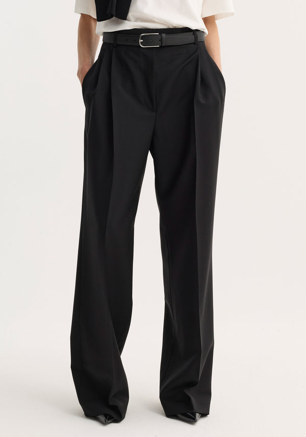 Model wearing mid-waist tailored trousers in black, featuring double pleats, wide legs, and slim belt, crafted from virgin wool-mohair blend for structure and breathability.