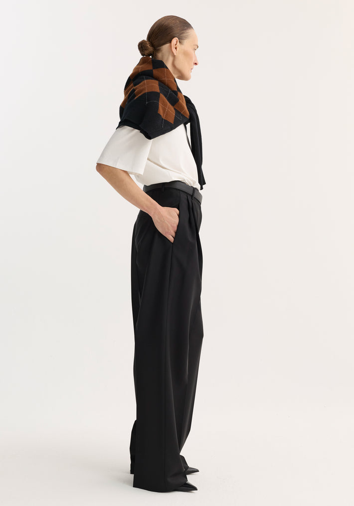 Person in profile wearing mid-waist tailored trousers in black, featuring a wide leg and double pleats, styled with a casual sweater over shoulders.