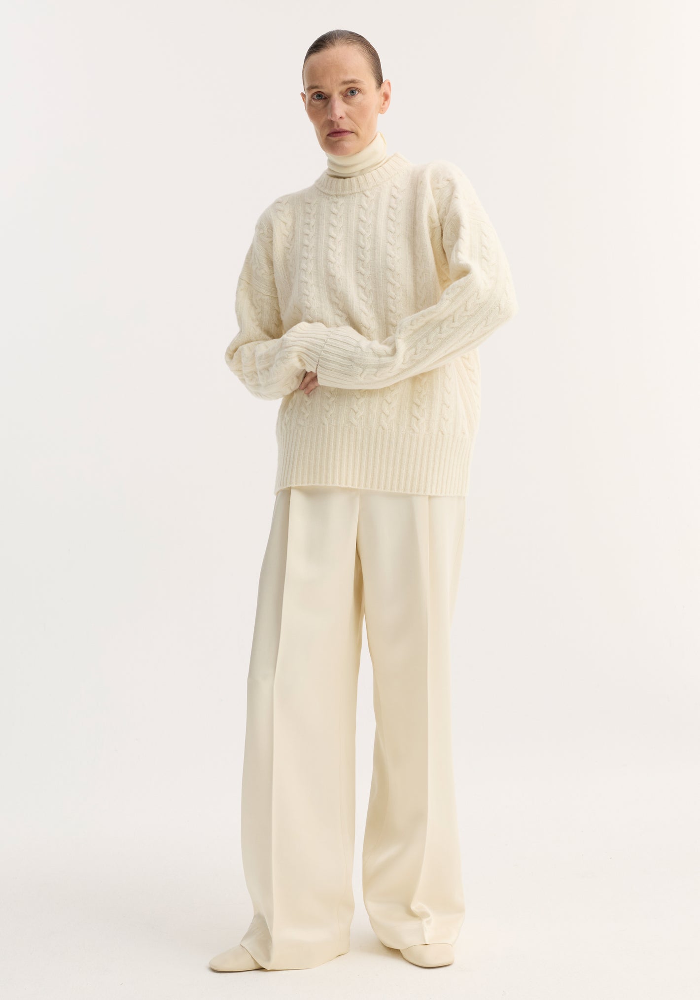 Model wearing Relaxed drawstring satin trousers in cream, featuring a long wide-leg silhouette, drawstring waistband, and back welt pockets, paired with a cable-knit sweater.