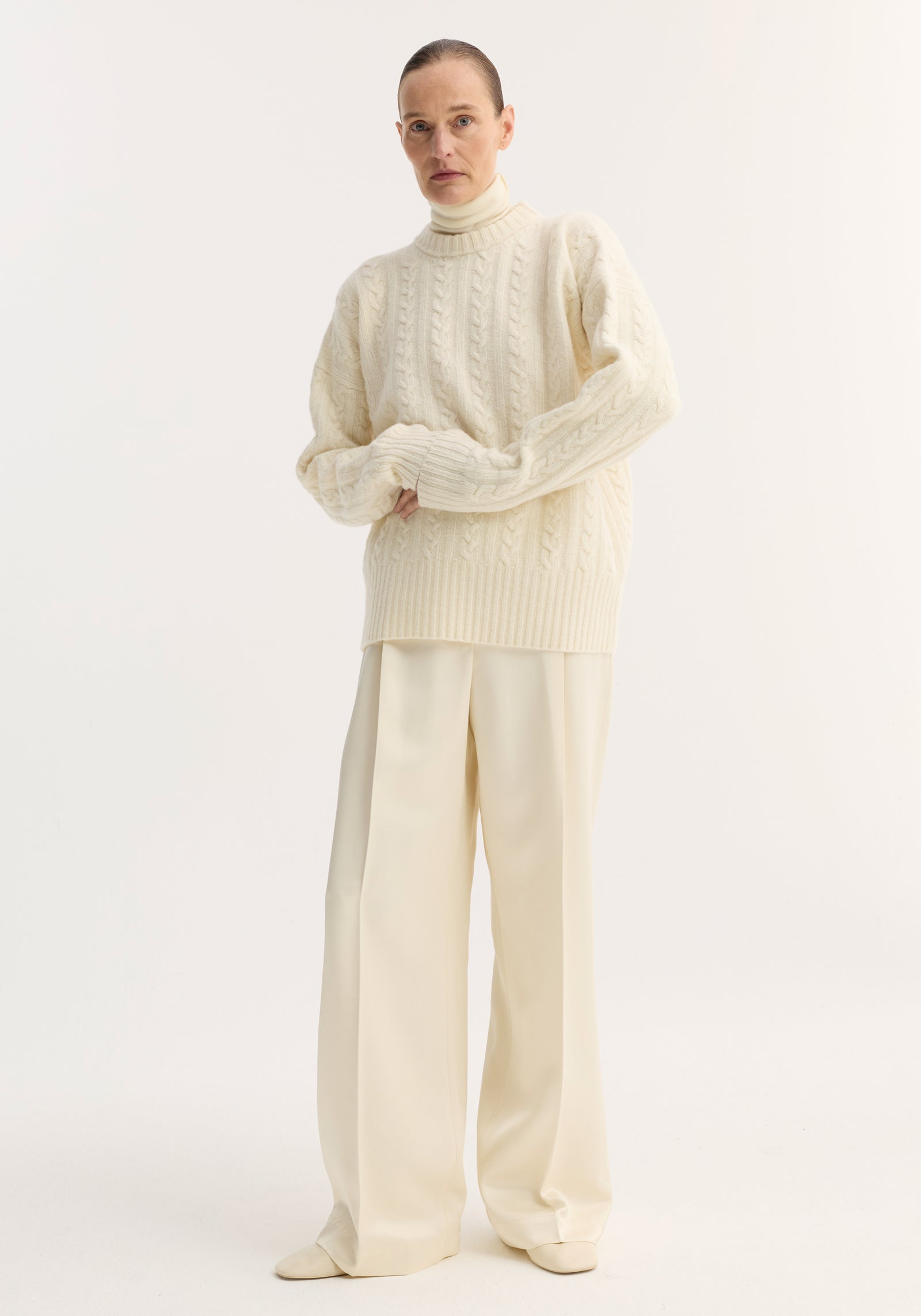 Model wearing Relaxed drawstring satin trousers in cream, featuring a long wide-leg silhouette, drawstring waistband, and back welt pockets, paired with a cable-knit sweater.