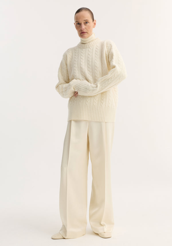 Model wearing Relaxed drawstring satin trousers in cream, featuring a long wide-leg silhouette, drawstring waistband, and back welt pockets, paired with a cable-knit sweater.