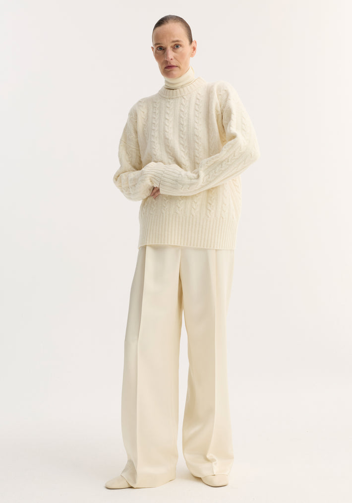 Model wearing Relaxed drawstring satin trousers in cream, featuring a long wide-leg silhouette, drawstring waistband, and back welt pockets, paired with a cable-knit sweater.