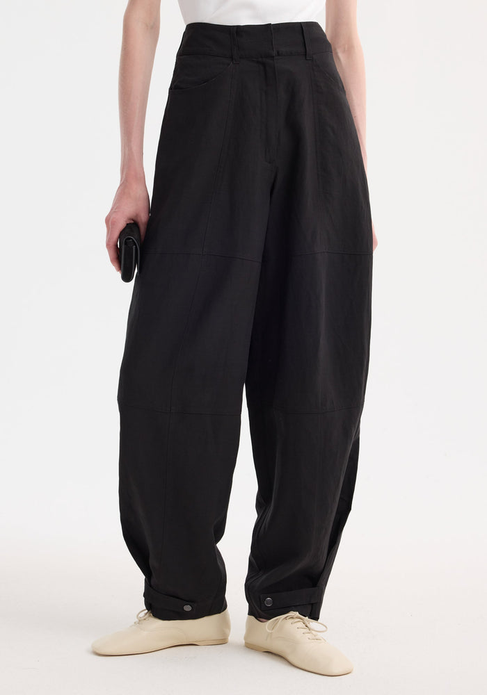 Parachute trousers in black, high-waisted with wide legs, tapering at ankles with snap-tabs. Paired with cream lace-up shoes and a black clutch.
