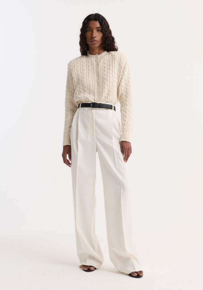 Woman wearing Mid-waist tailored trousers in off white with a tailored waistband, double pleats, and wide-leg design, paired with a cream cable-knit sweater.