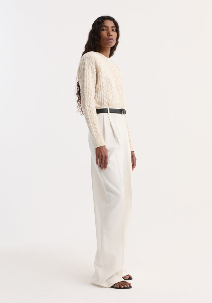 Woman models Mid-waist tailored trousers in off white, showcasing high-waisted, wide-leg design with pleats and black belt, paired with cream cardigan.