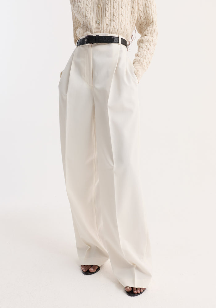 Mid-waist tailored trousers in off white; features high-waist, wide-leg fit, sharp pleats, and side pockets, paired with a black belt and heeled sandals.