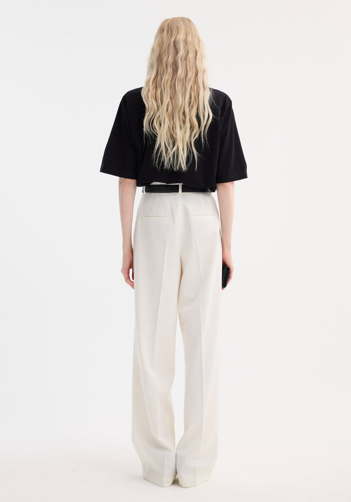 Person with long blonde hair wears mid-waist tailored trousers in off white, featuring a wide leg and double pleats, holding a black clutch.