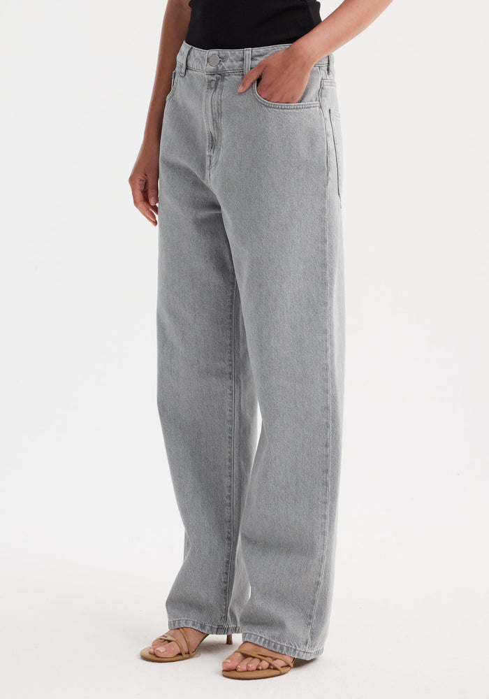 Model wearing Signature baggy fit denim in washed grey, wide-leg style, natural waist fit, with front pockets and tan strappy sandals, studio setting.