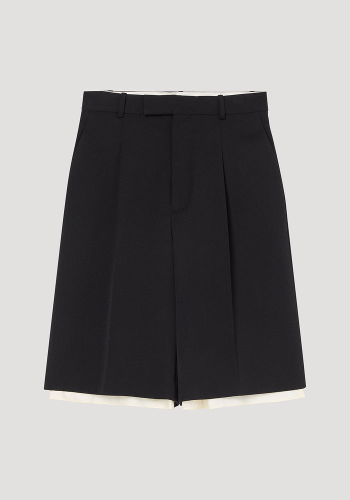 Contrast extended lining shorts in black, featuring high-waisted design, skirt-like front, belt loops, side pockets, and a subtle cream lining peeking beneath the hem.