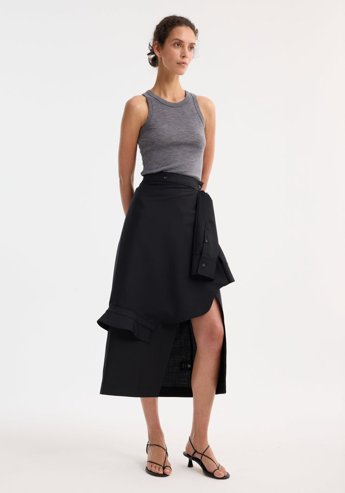Person wearing a Shirt wrap skirt in black, featuring an asymmetrical wrap, front slit, and deconstructed sleeves, paired with a heather-gray tank top and black sandals.