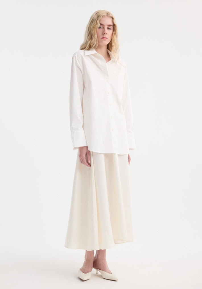Model in Poplin silk combined gilet-dress with oversized shirt and flowing skirt, showcasing a minimalist look with a sculptural, deconstructed design and buttoned wrap belt.
