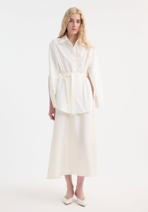 Model in Poplin silk combined gilet-dress with oversized shirt and flowing skirt, showcasing a minimalist look with a sculptural, deconstructed design and buttoned wrap belt.