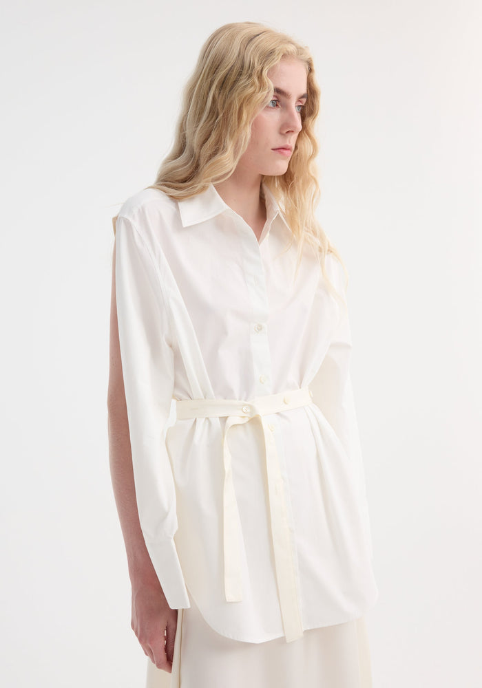Poplin silk combined gilet-dress in optic white on model, featuring a deconstructed style with open armholes, buttoned wrap belt, and layered cotton-silk design.