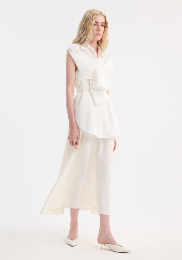 Model wearing the Poplin silk combined gilet-dress in optic white, featuring a deconstructed design with open armholes, buttoned wrap belt, and fluid silhouette.