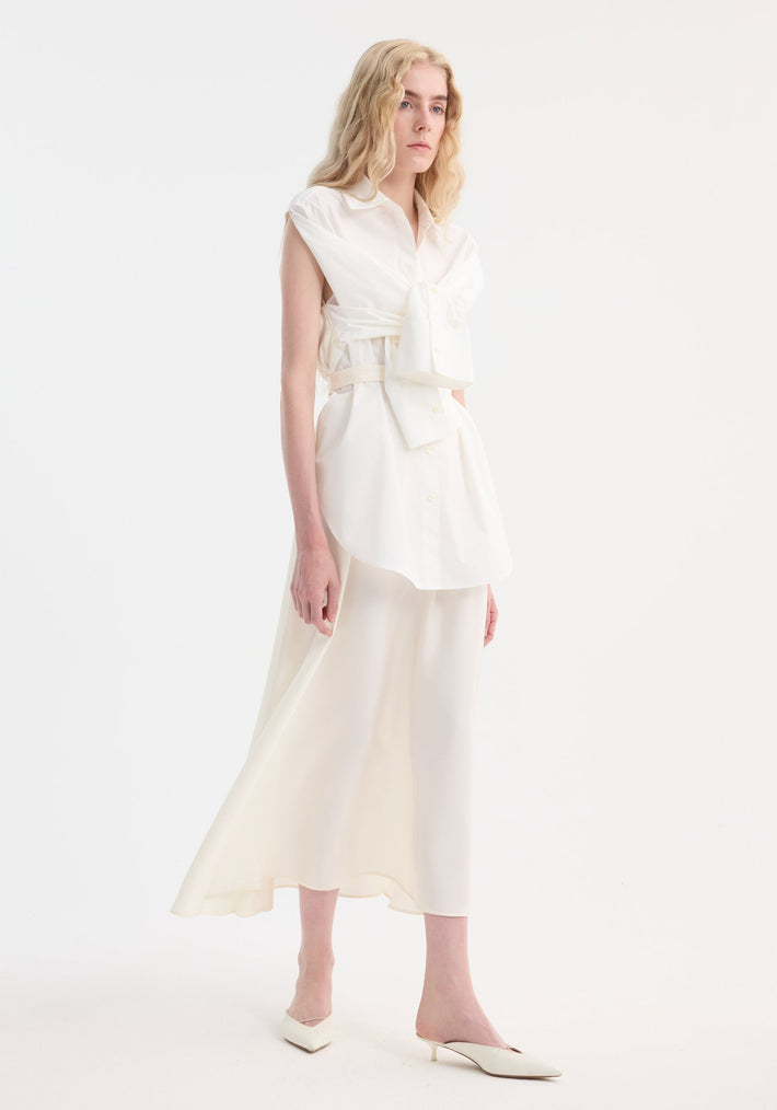 Model wearing the Poplin silk combined gilet-dress in optic white, featuring a deconstructed design with open armholes, buttoned wrap belt, and fluid silhouette.