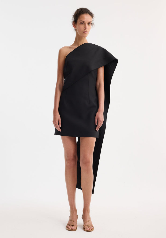 Short sculptural sash dress in black, featuring a one-shoulder design with an asymmetric draped sash, fitted silhouette, and beige strappy sandals.