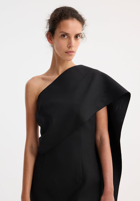 Short sculptural sash dress in black, featuring a one-shoulder design with an asymmetric draped sash, fitted silhouette, and beige strappy sandals.