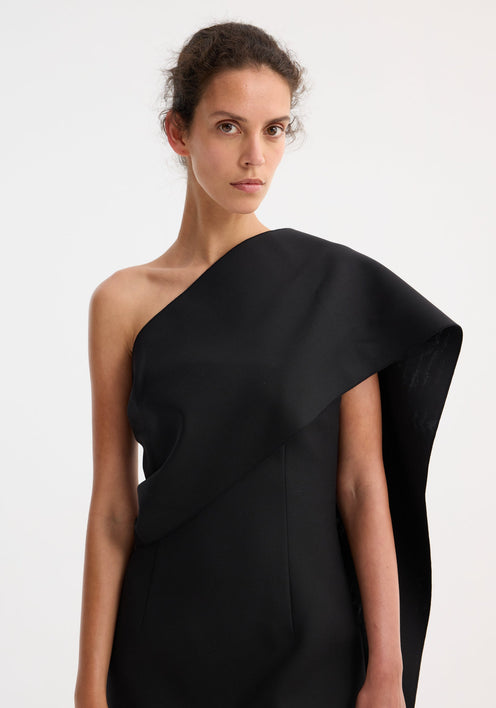 Short sculptural sash dress in black, featuring a one-shoulder design with an asymmetric draped sash, fitted silhouette, and beige strappy sandals.