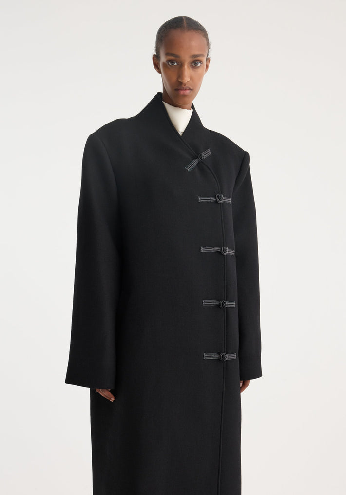 Model wearing Mandarin wool coat with asymmetric mandarin closures, long length, and minimal design, crafted from 100% virgin wool against a plain background.