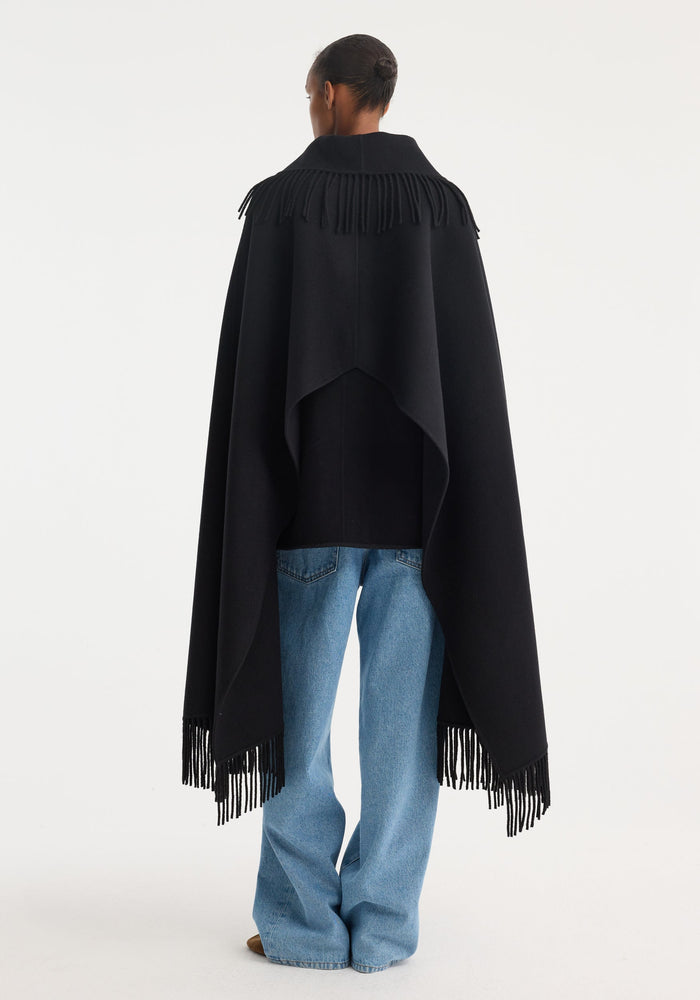 Person wearing a Fringe poncho double-faced wool in black, showcasing its wide collar and fringed edges, paired with light-wash jeans and brown shoes.