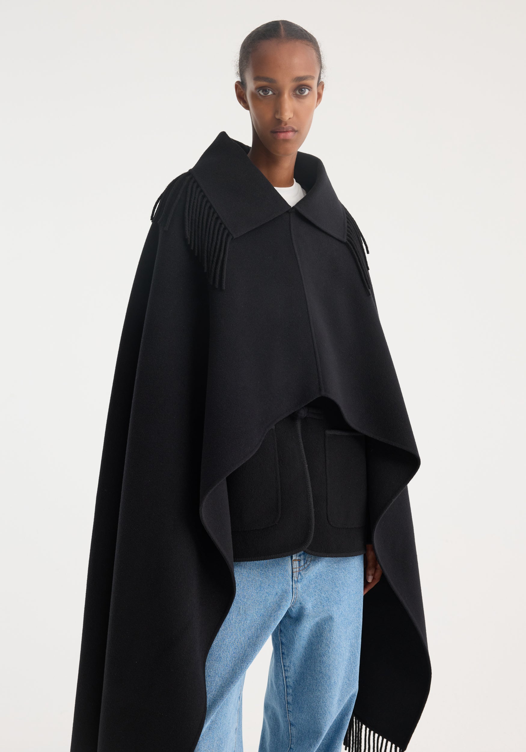 Model wearing a black Fringe poncho double-faced wool | black with wide collar and fringe details on the shoulders, paired with light blue jeans against a white background.