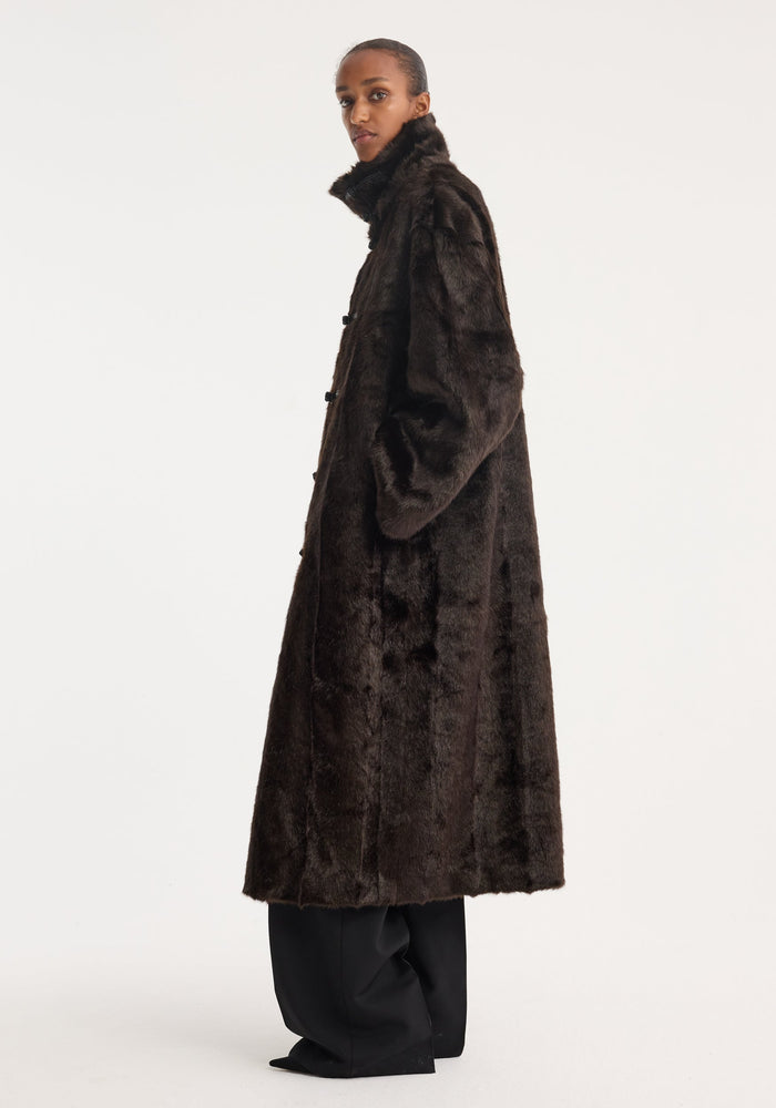 Model in profile wearing a Mandarin faux fur coat in dark brown, featuring mandarin front closures and a stand-up collar, with visible wide-leg trousers and pointed-toe shoes.