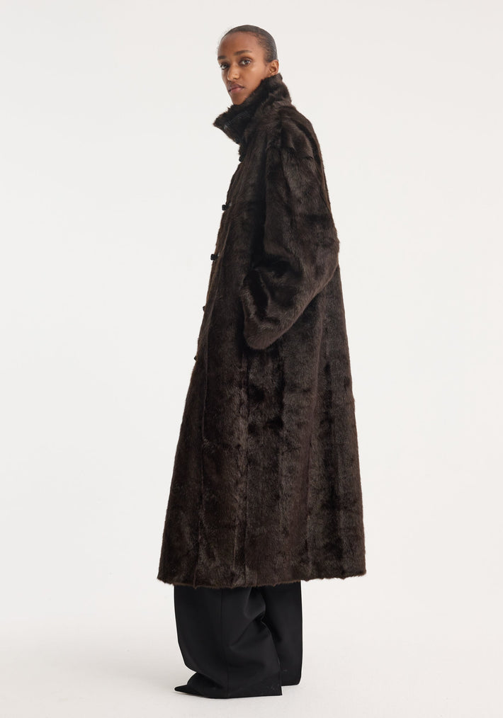 Model in profile wearing a Mandarin faux fur coat in dark brown, featuring mandarin front closures and a stand-up collar, with visible wide-leg trousers and pointed-toe shoes.