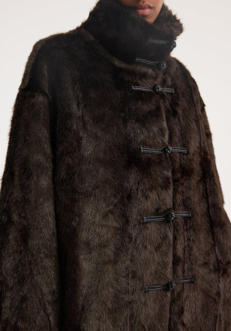 Model wears a Mandarin faux fur coat in dark brown, featuring toggle closures and a stand-up collar, paired with wide-leg black trousers, exuding a chic, sophisticated look.