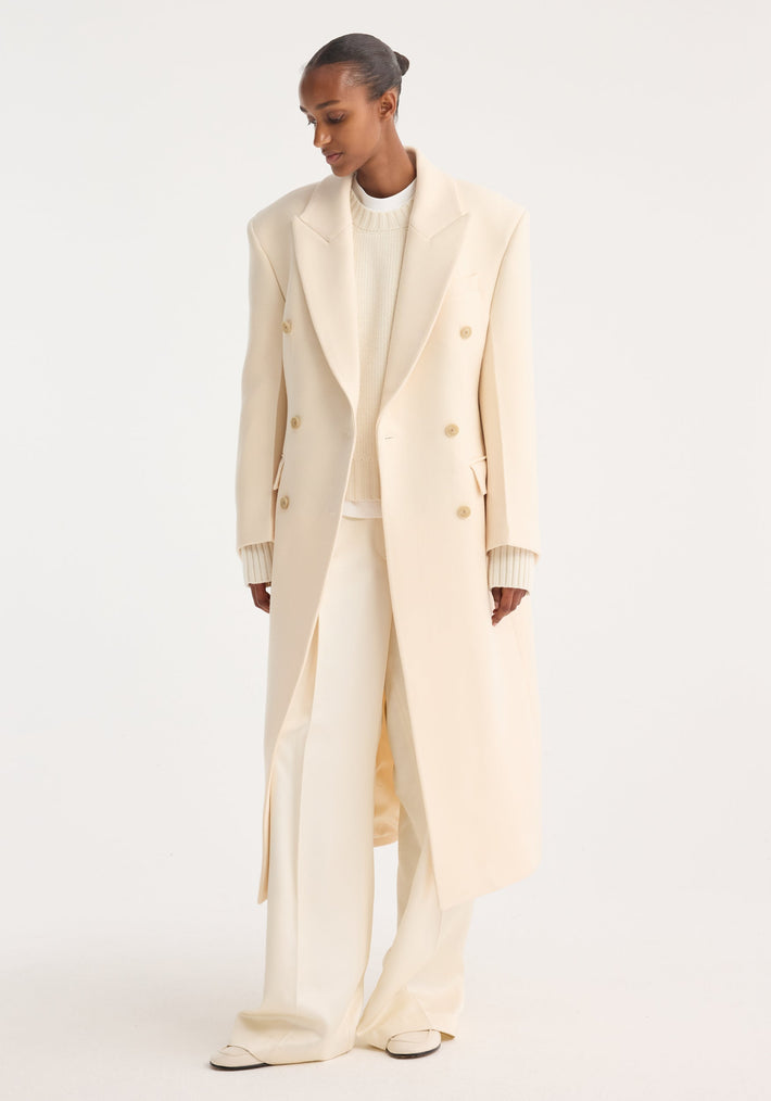Hourglass wool coat in off white, featuring double-breasted design and wide lapels, worn over a chunky knit sweater and wide-leg trousers, showcasing minimalist elegance.