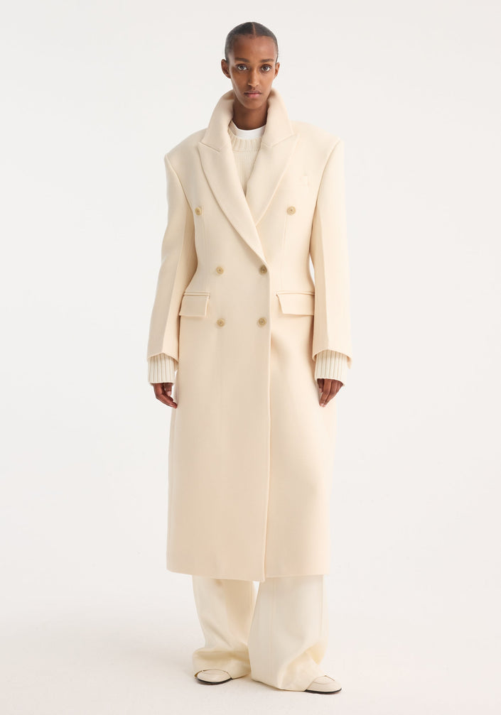 Model wearing Hourglass wool coat, off white, featuring a double-breasted design and strong shoulders, paired with matching trousers and loafers for a minimalist look.