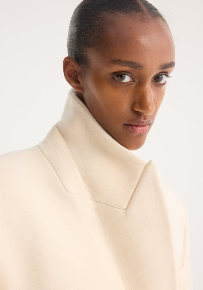 Hourglass wool coat in off white, featuring a high collar and structured design, worn by a model with slicked-back hair, highlighting the coat's elegant hourglass silhouette.