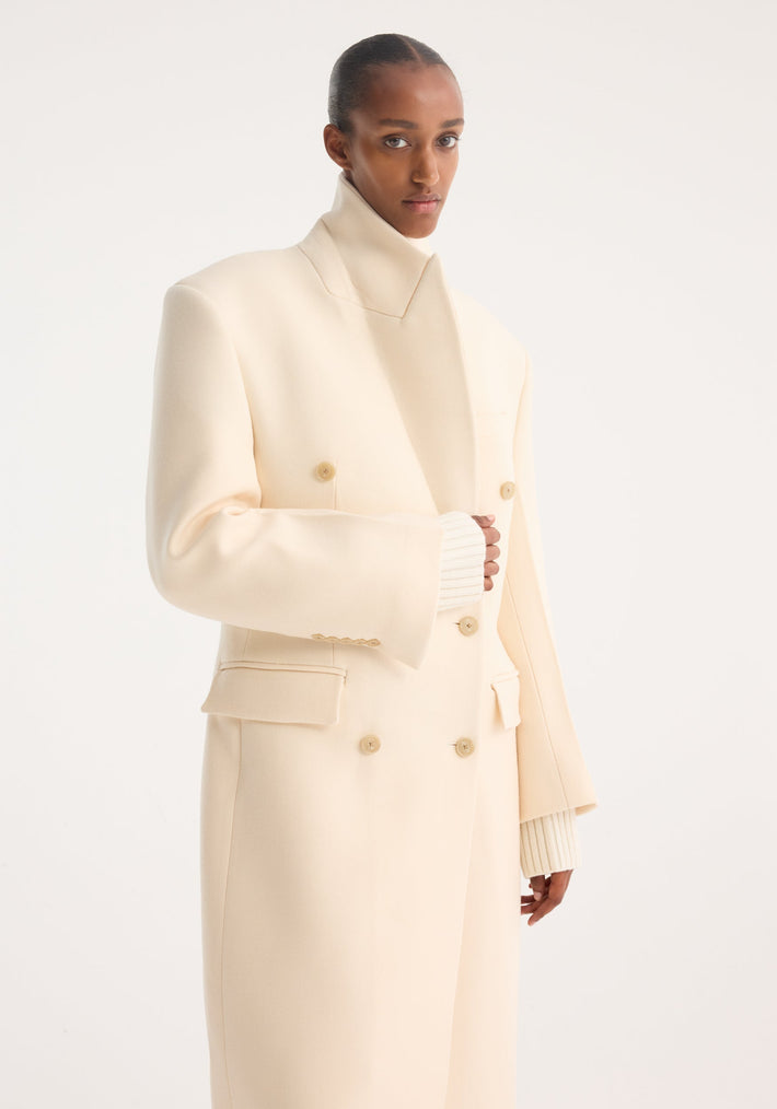 Person wearing an Hourglass wool coat in off white, featuring a long, double-breasted design with a high stand collar, showcasing tailored elegance and ribbed knit cuffs.