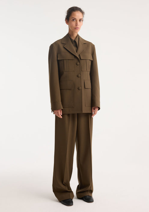 Model in Safari blazer-jacket, forest, featuring boxy notched collar, four patch pockets, wide-leg trousers, paired with pointed-toe loafers, in a studio setting.