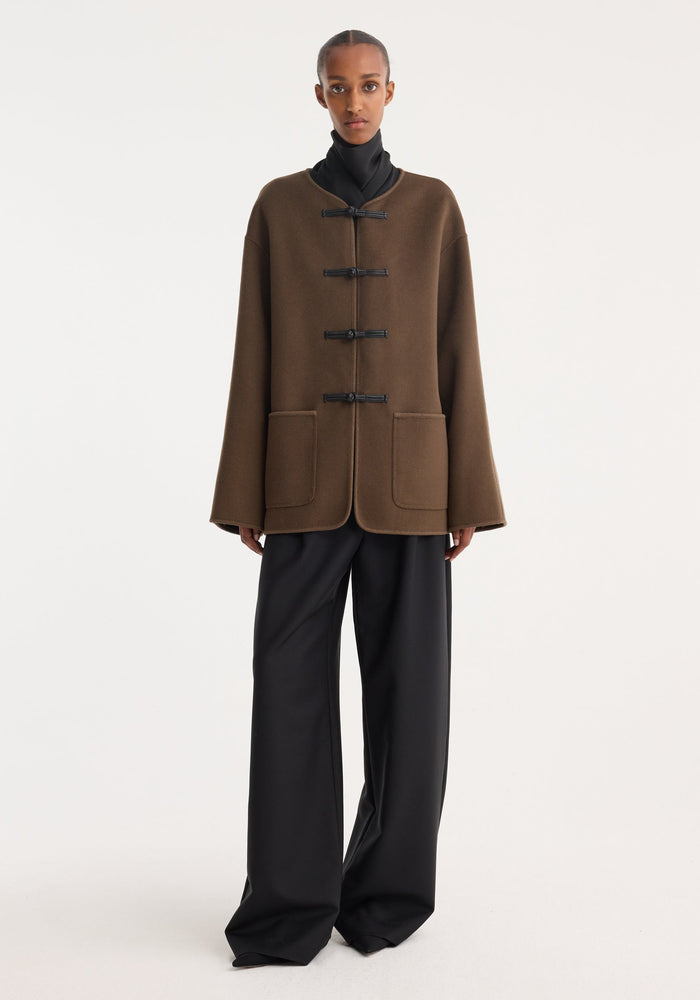 Model in Mandarin double-faced wool jacket | taupe, featuring black toggle closures, patch pockets, with embroidered edges, styled over a high-neck top and wide-leg trousers.